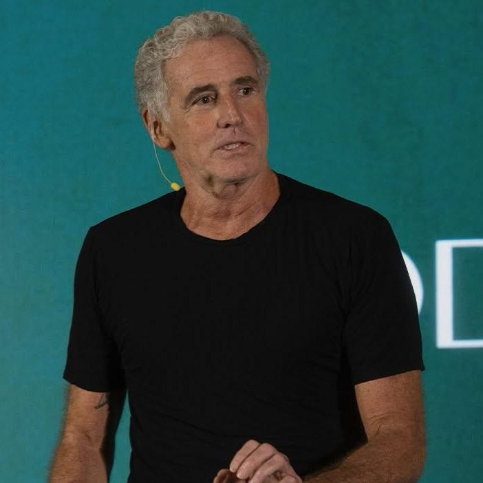 Marcelo Politi em palestra, de camiseta preta, diante de um telão